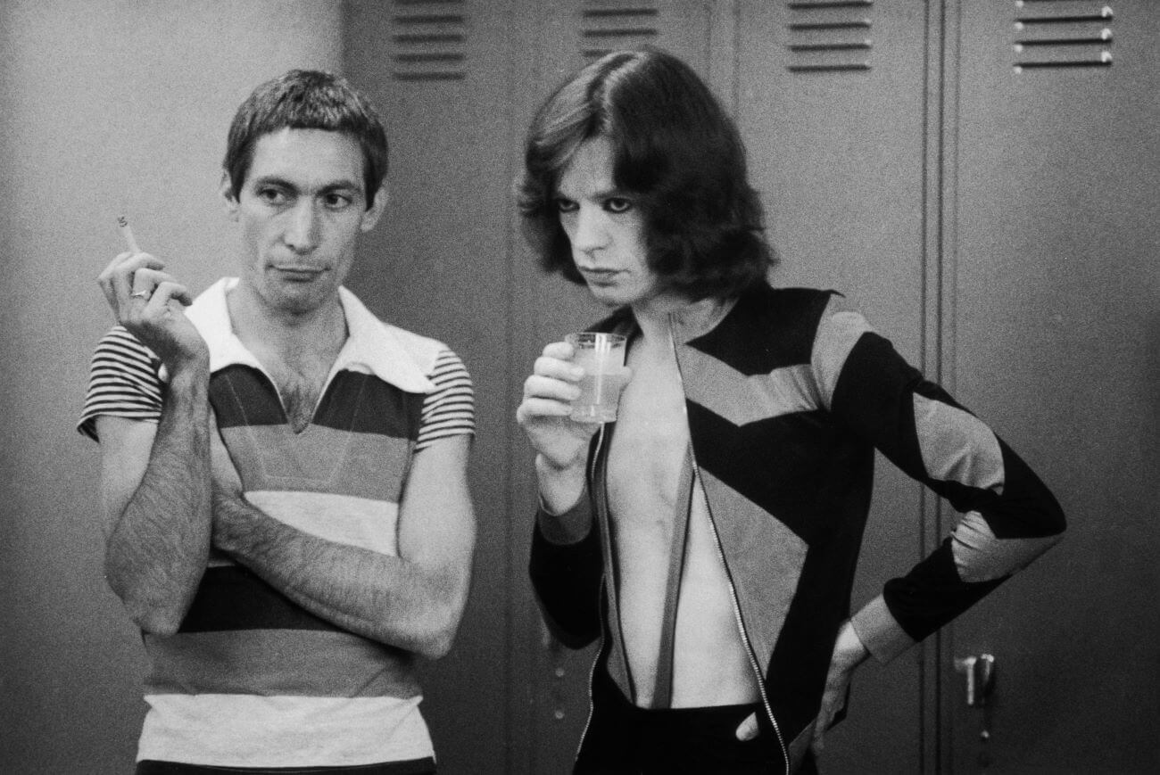 A black and white picture of Charlie Watts holding a cigarette and Mick Jagger holding a small glass. They stand in front of a row of lockers.