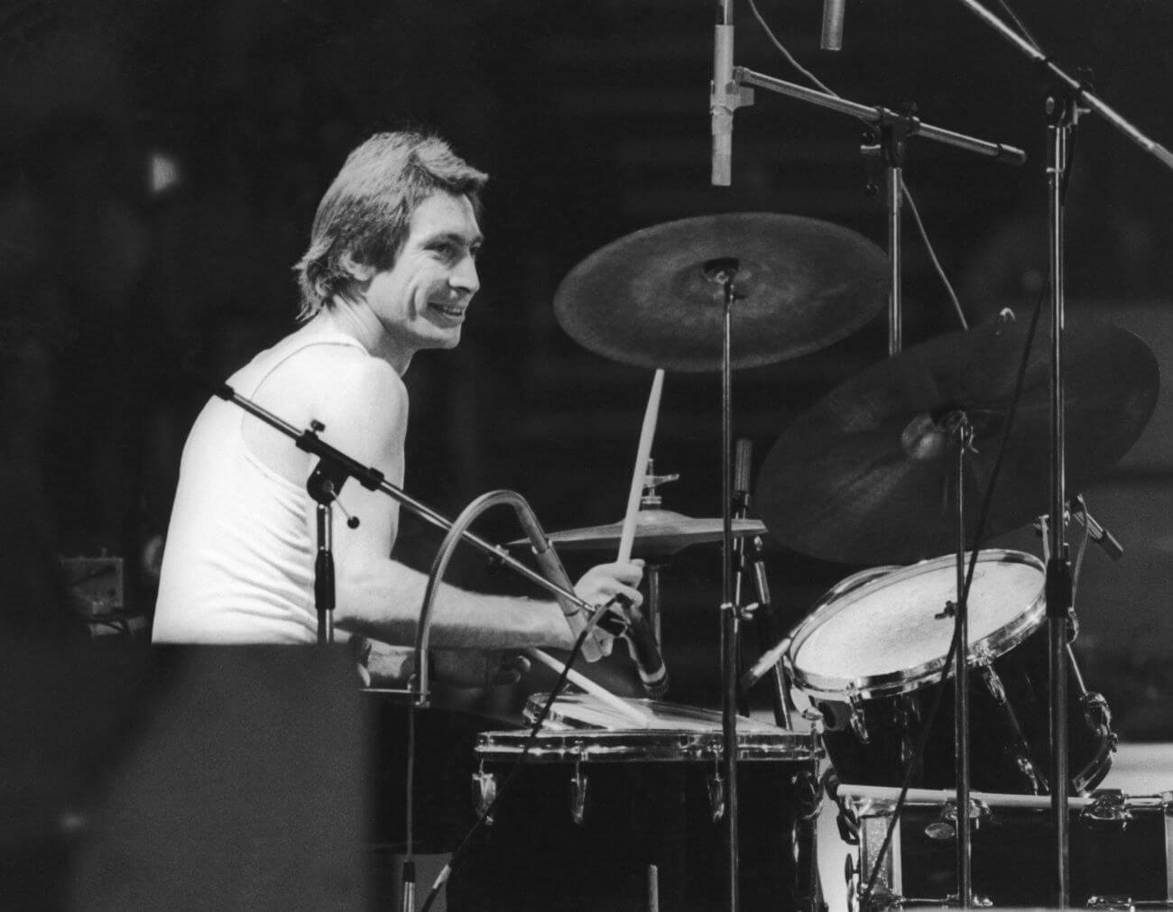 A black and white picture of Rolling Stones drummer Charlie Watts wearing a tank top and sitting at a drum set.