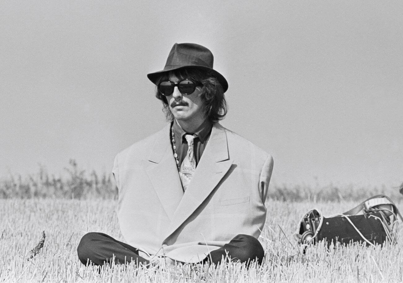 A black and white picture of George Harrison wearing an oversized coat, sunglasses, and hat and sitting in a field.