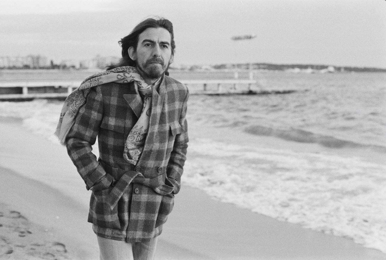 A black and white picture of George Harrison wearing a coat and scarf and walking on the beach.