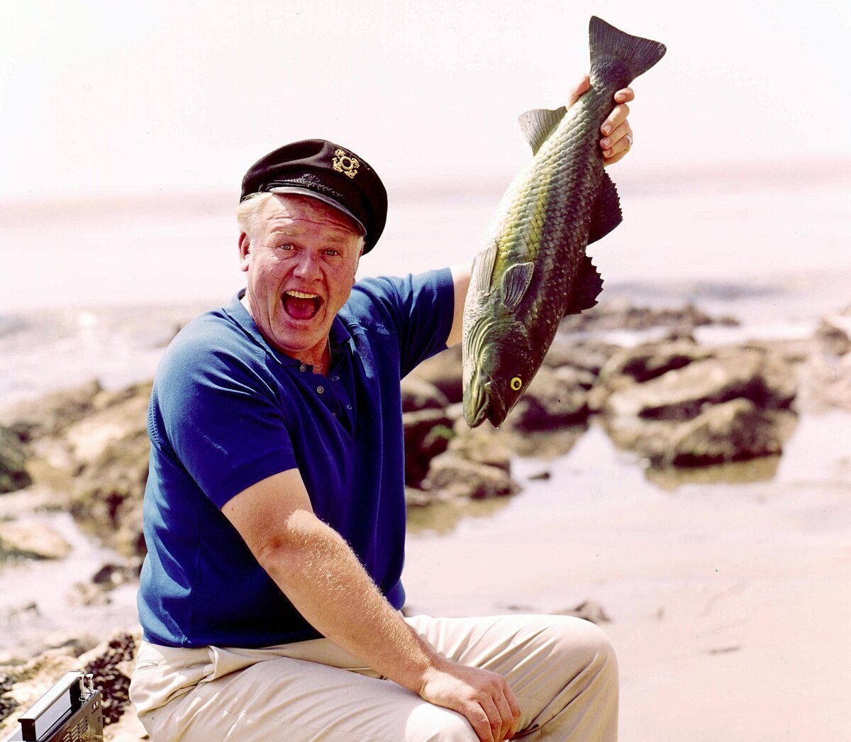 Alan Hale Jr. holds a fish while sitting on the beach as The Skipper from 'Gilligan's Island'