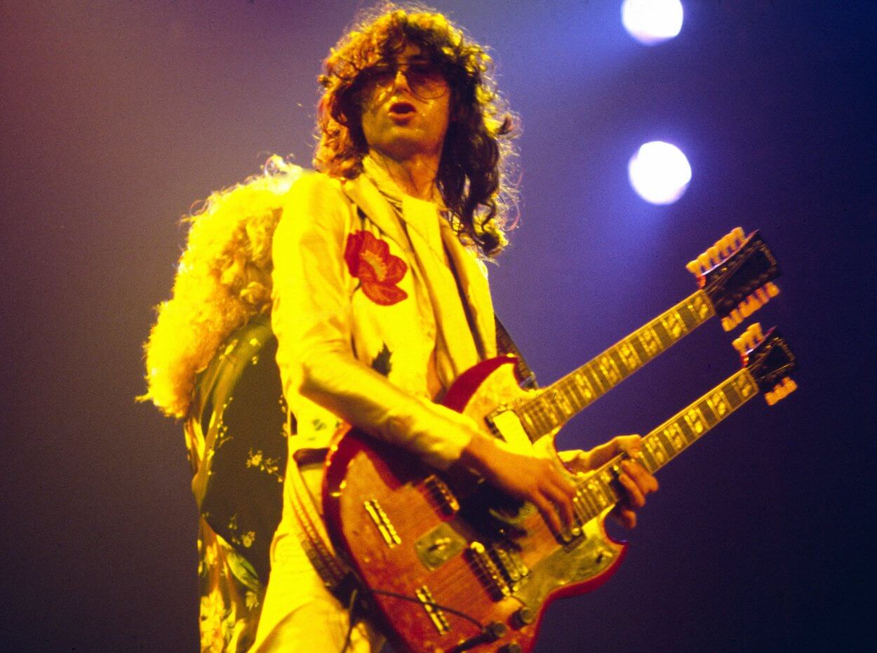 Jimmy Page wearing a white suit with a red poppy design while playing a double-necked guitar in 1977.