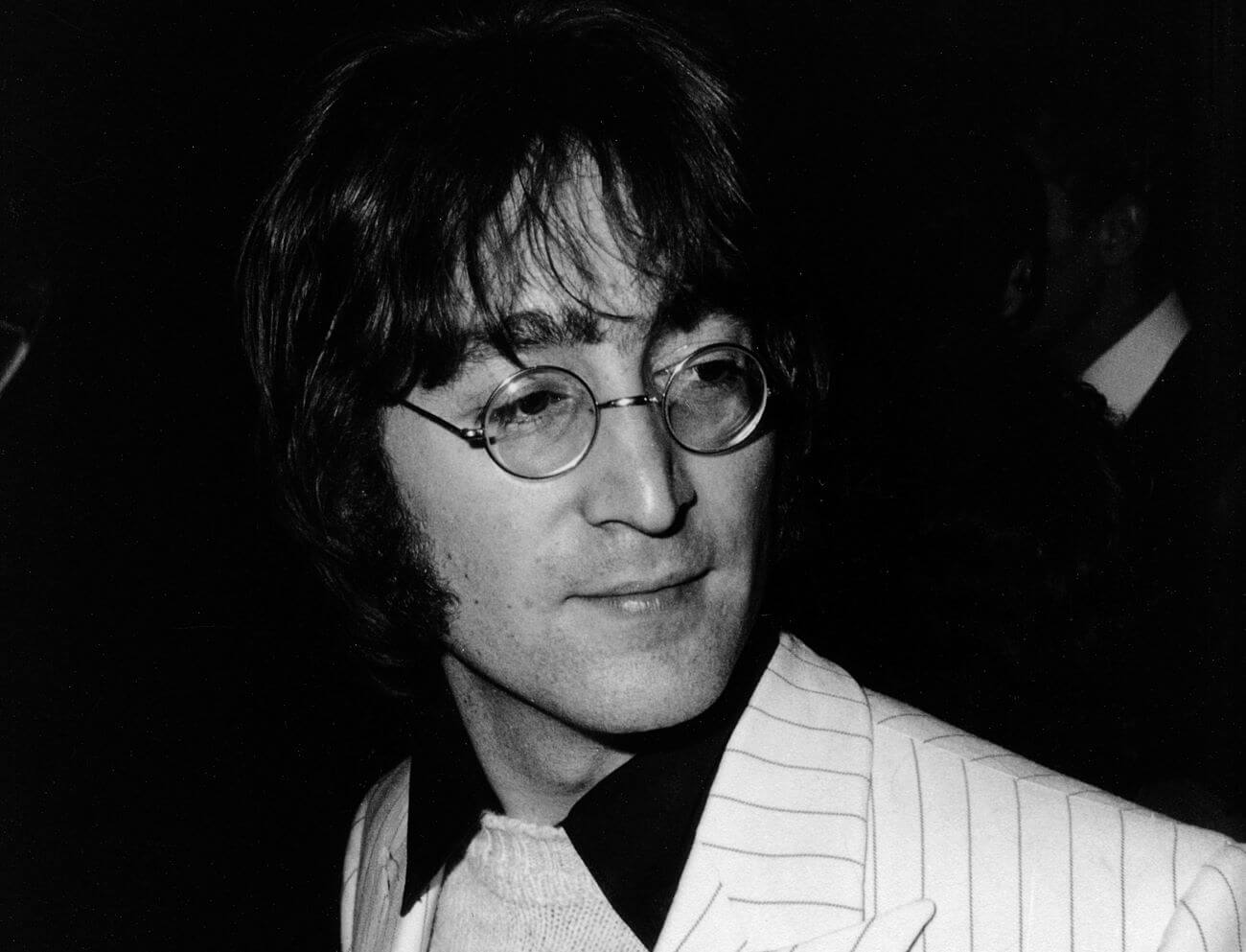 A black and white picture of John Lennon wearing round glasses and a pinstripe coat.