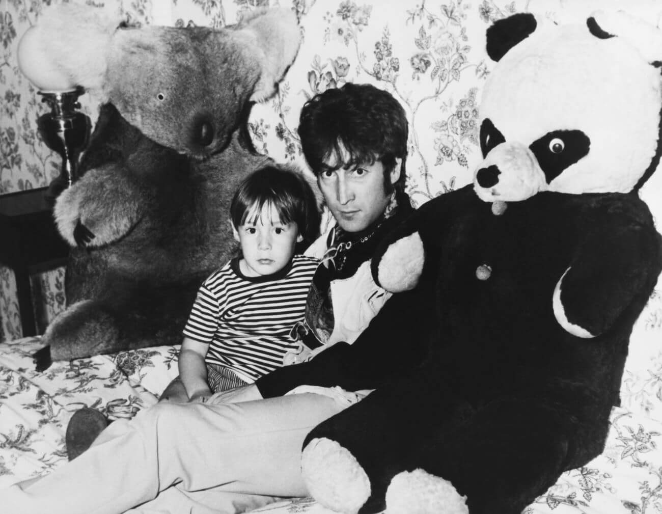 A black and white picture of Julian Lennon sitting with John Lennon sitting on a bed between two large stuffed bears.