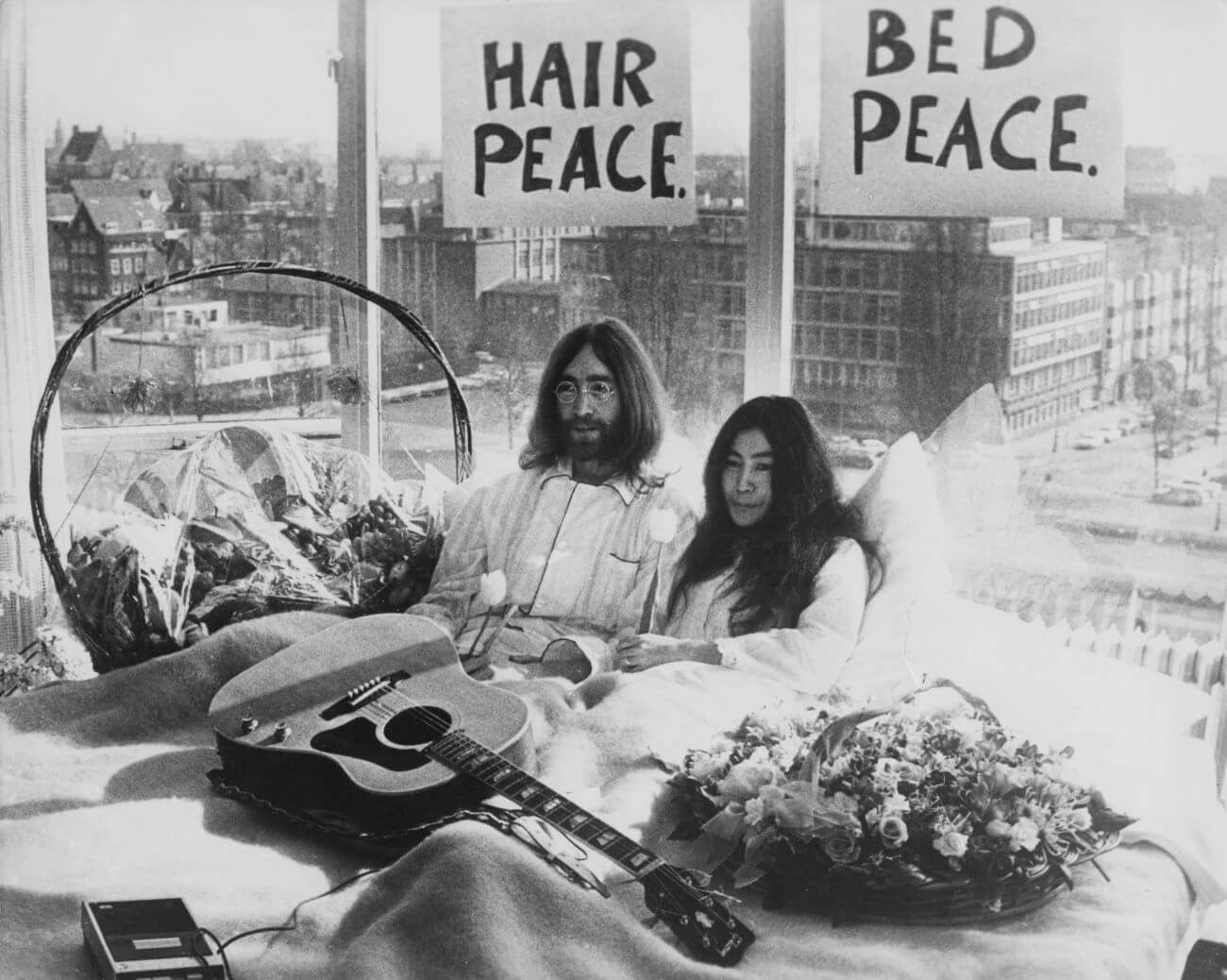 John Lennon and Yoko Ono sit in bed with baskets of flowers and a guitar. Signs that say 