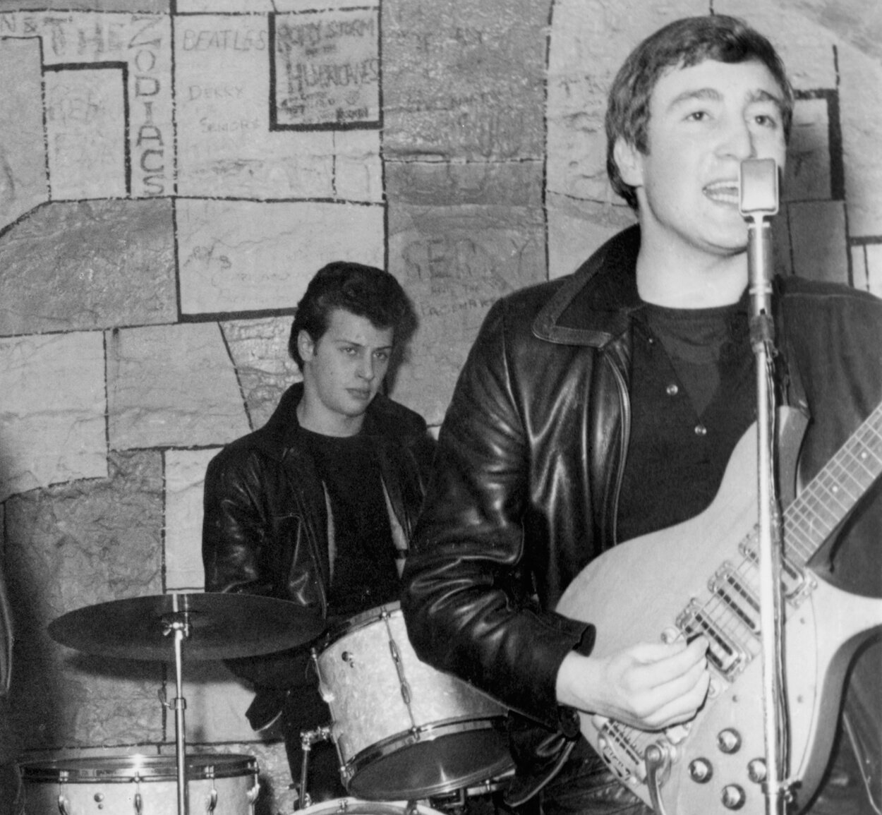 Pete Best (left, background) drumming while John Lennon sings and plays guitar at a 1961 Beatles concert.