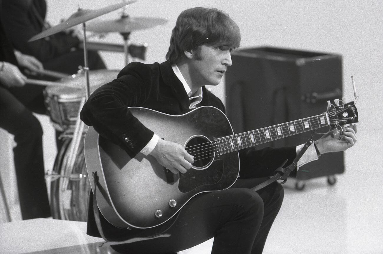 A black and white picture of John Lennon sitting in front of a drum set with a guitar.