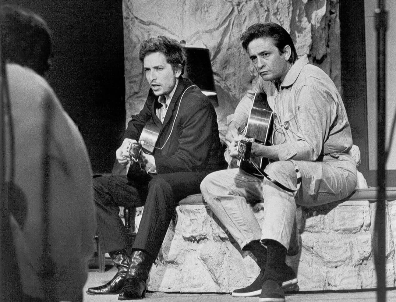 A black and white picture of Bob Dylan and Johnny Cash playing guitars and sitting on a stone bench.