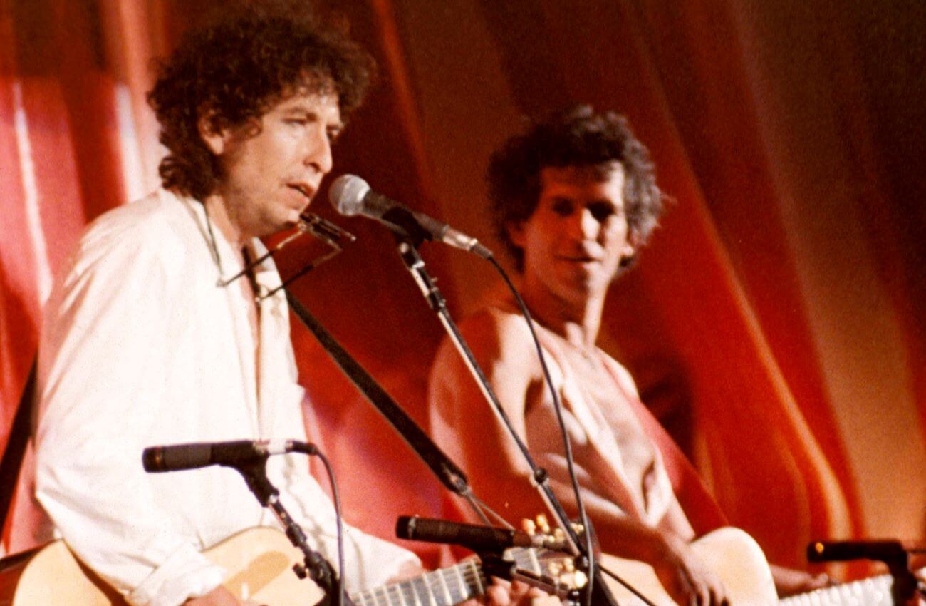 Bob Dylan sings into a microphone while Keith Richards watches. Both hold acoustic guitars.