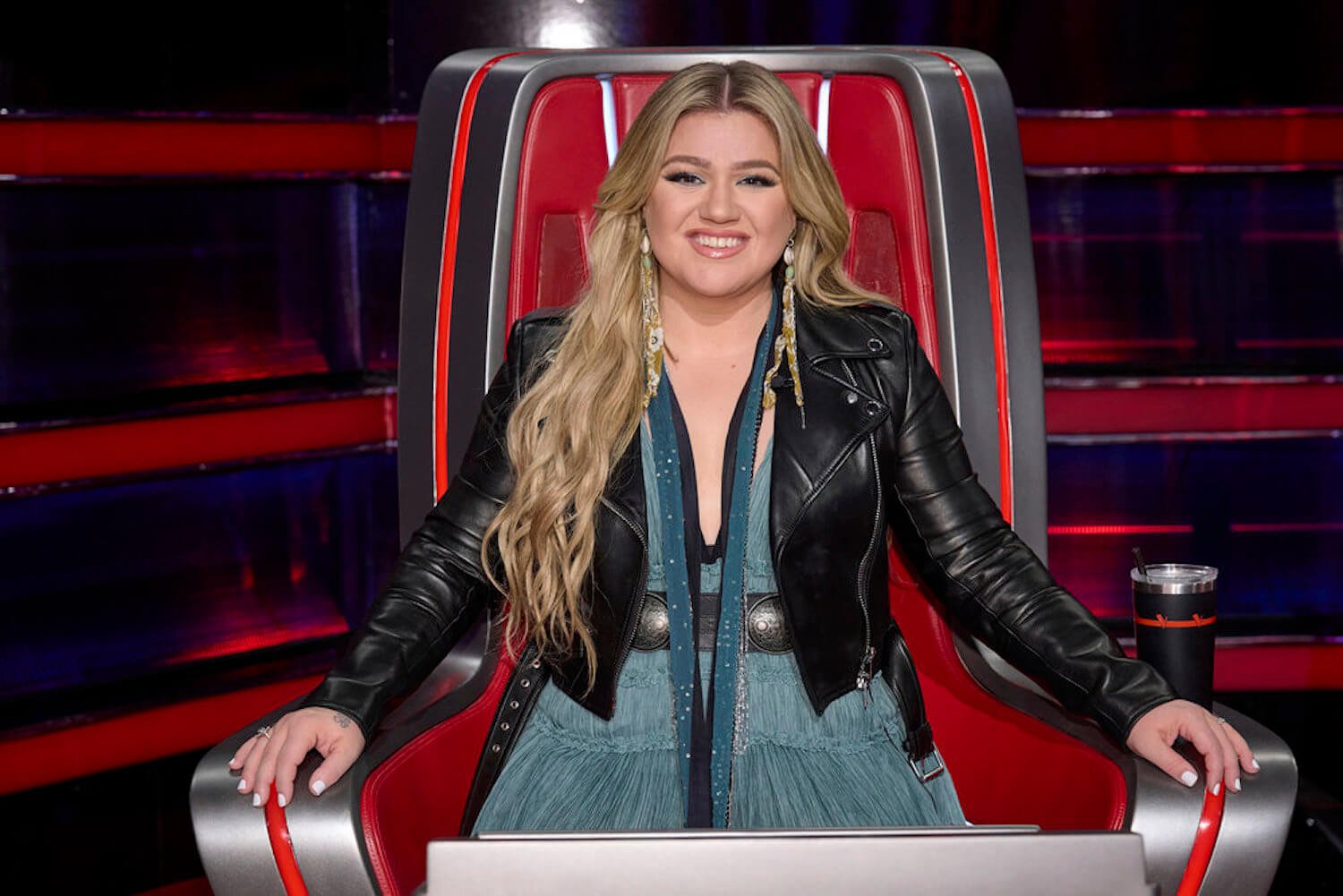 Kelly Clarkson smiling and sitting in a red chair on 'The Voice'