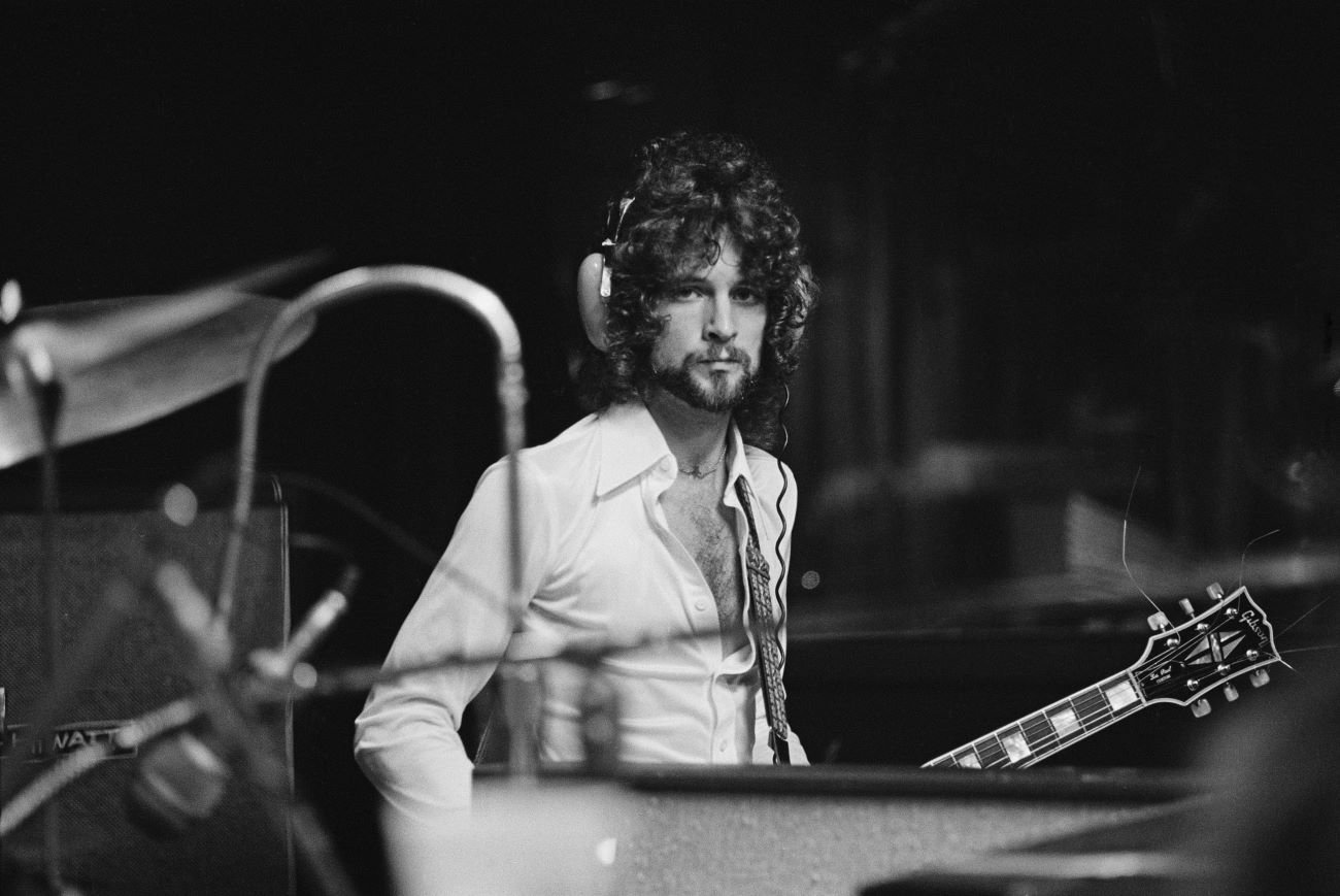 A black and white picture of Lindsey Buckingham of Fleetwood Mac wearing headphones and holding a guitar.