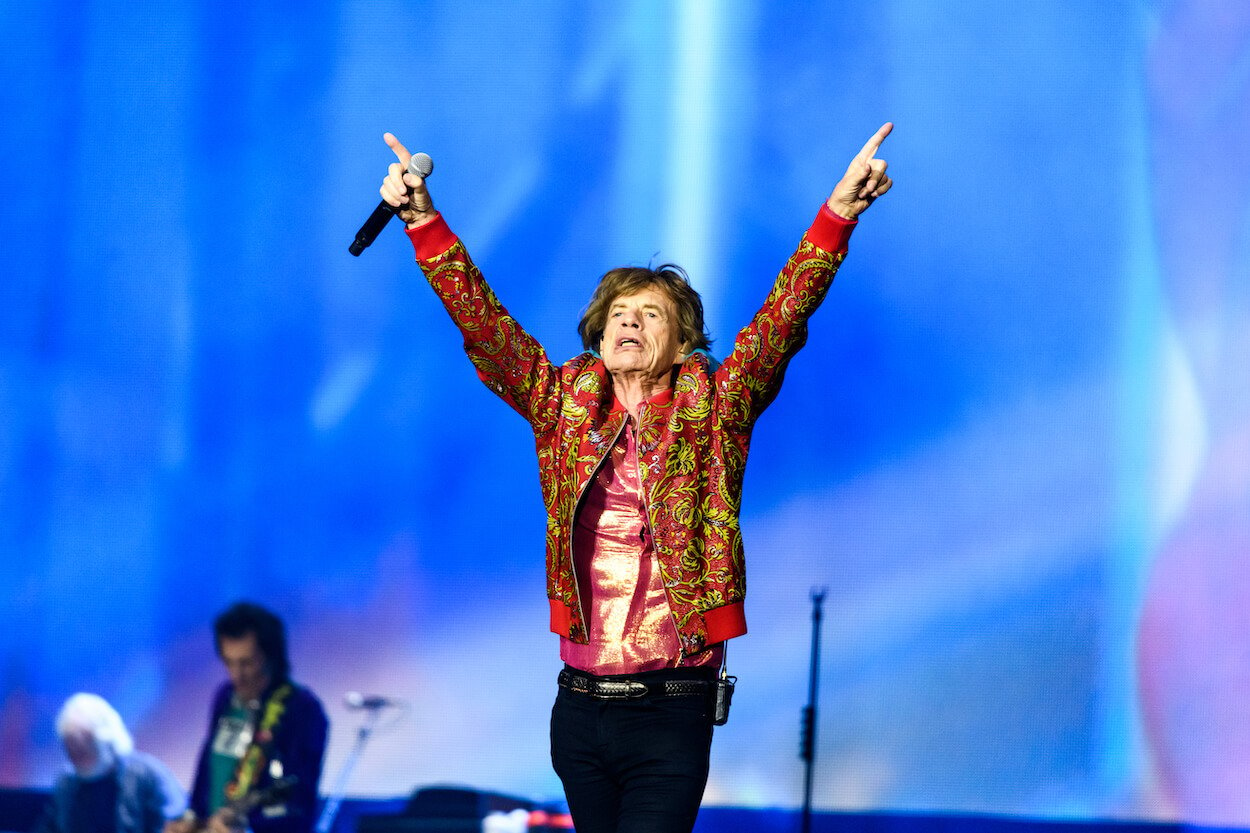 Rolling Stones frontman Mick Jagger wearing a red, patterned jacket while holding a microphone and pointing at the sky during a concert.