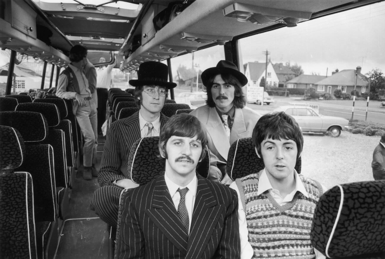 A black and white picture of John Lennon, George Harrison, Ringo Starr, and Paul McCartney  sitting on a bus together on the set of the Beatles' film "Magical Mystery Tour."
