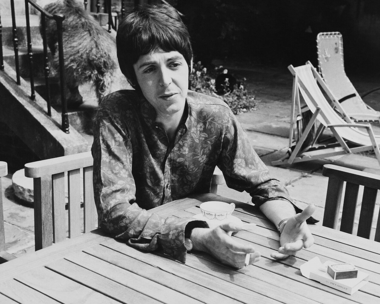 Beatles bassist Paul McCartney sitting at an outdoor table while wearing a floral-patterned shirt and smoking a cigarette in 1967.