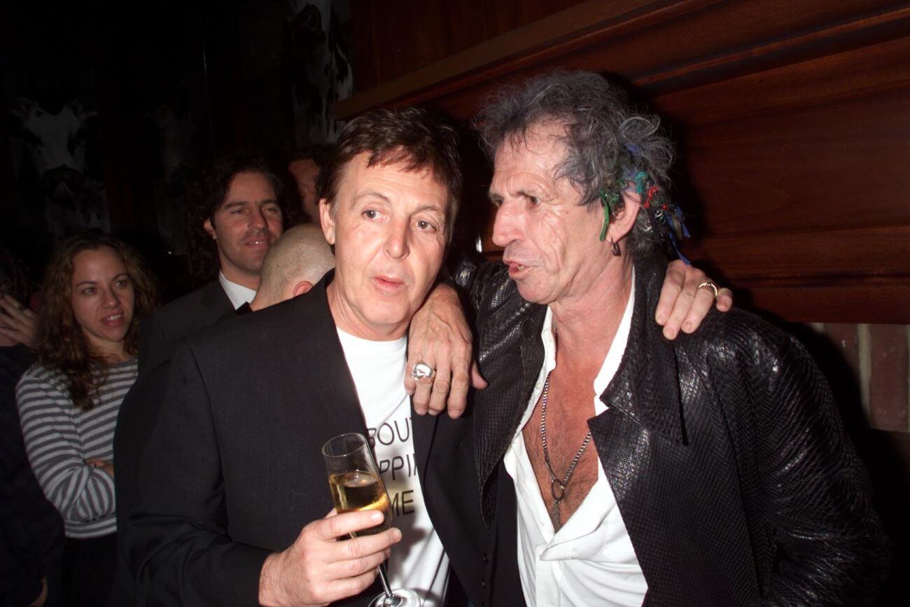 Paul McCartney and Keith Richards wear white shirts and black jackets and stand with their arms around each others' shoulders.
