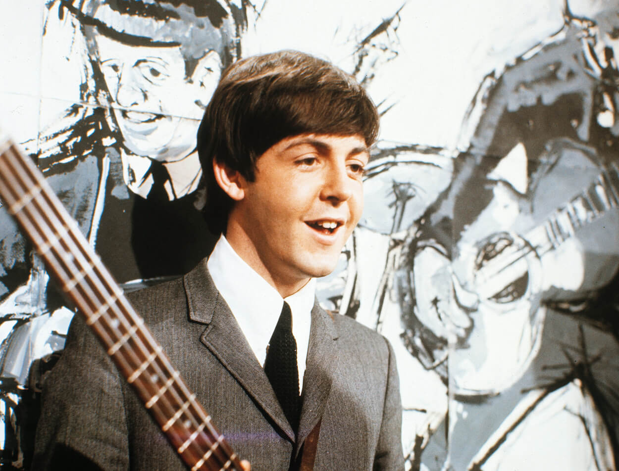 Paul McCartney holding his bass and smiling as he stands in front of a mural in 1962.