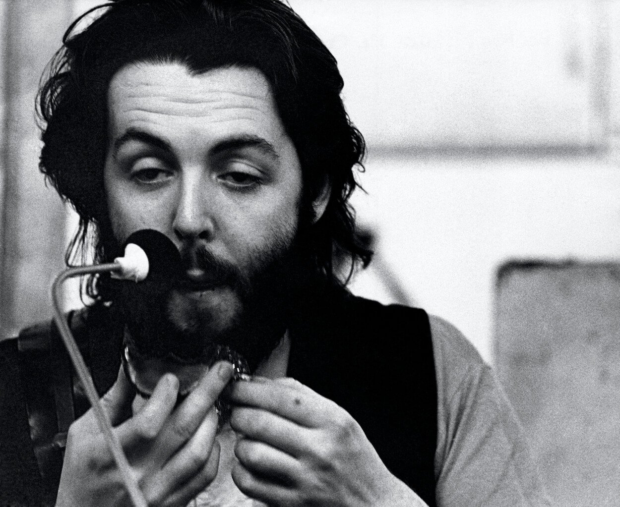 A bearded Paul McCartney wearing a dark vest over a shirt while speaking into a microphone circa 1970.