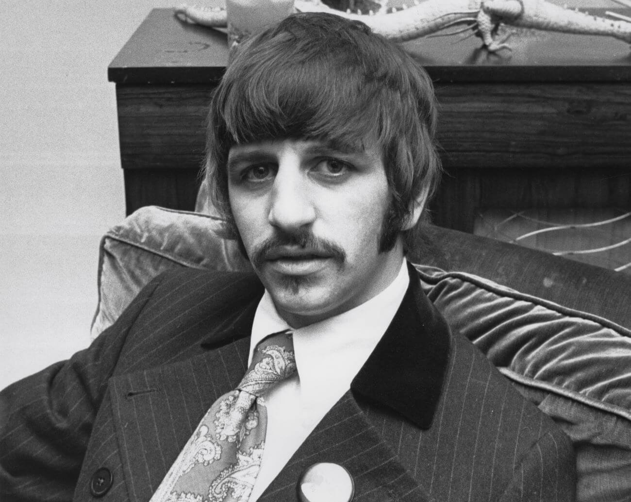 Ringo Starr, the drummer of The Beatles, wears a tie and sits in an arm chair.