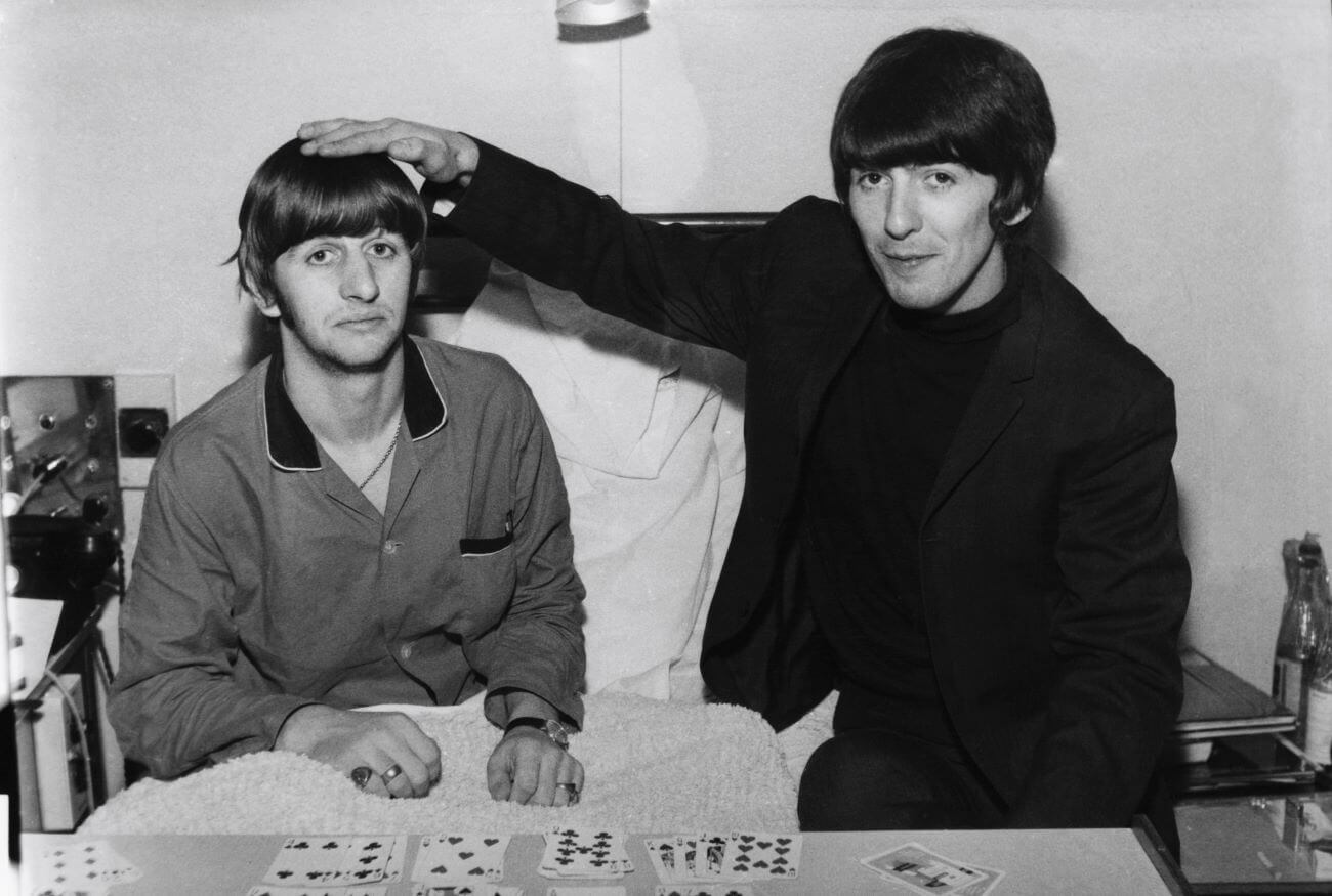 A black and white picture of Ringo Starr sitting in a hospital bed with cards in front of him and George Harrison at his bedside. Harrison rests his hand on Starr's head.