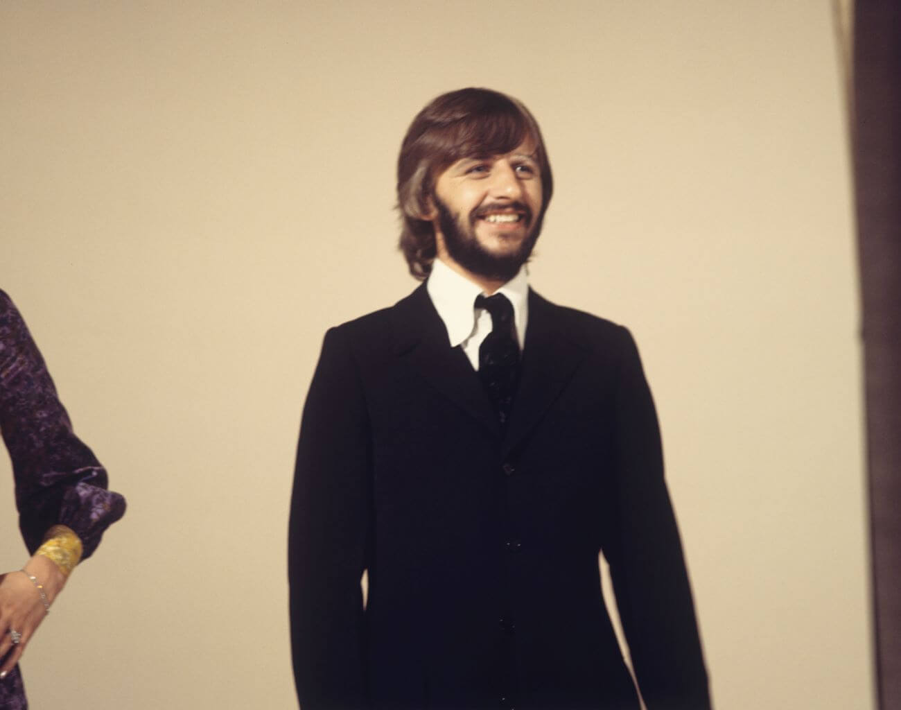 Ringo Starr wears a suit and stands in front of a tan background. 