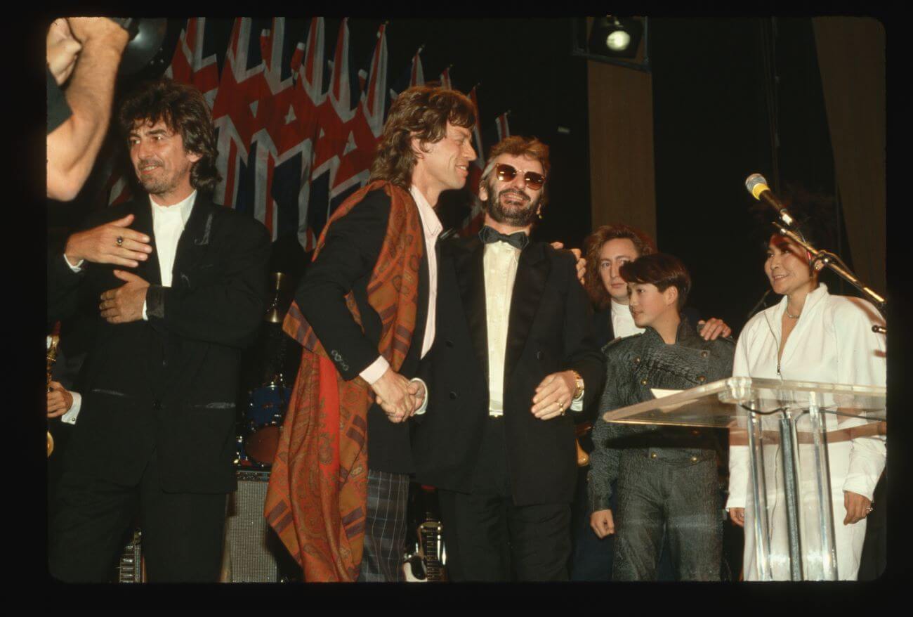 The Rolling Stones' Mick Jagger and Ringo Starr shake hands on a stage surrounded by George Harrison, Julian and Sean Lennon, and Yoko Ono.