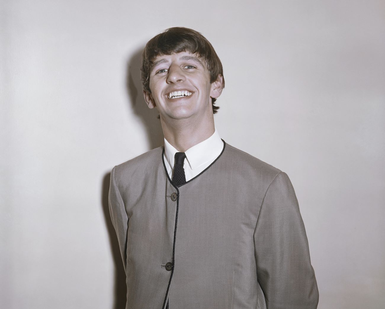 Ringo Starr wears a jacket and tie against a gray background. 