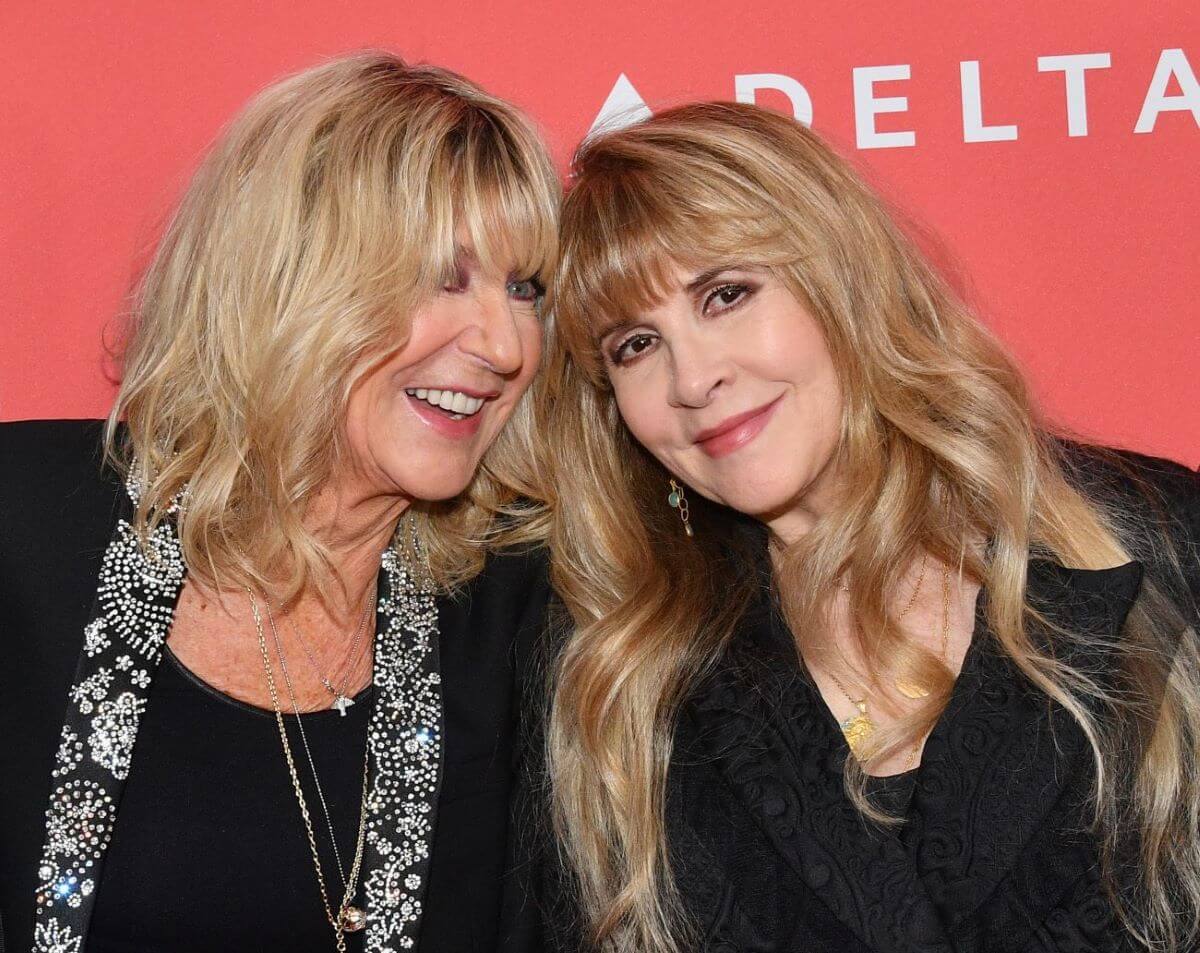 Christine McVie and Stevie Nicks wear black and smile together against a red background.