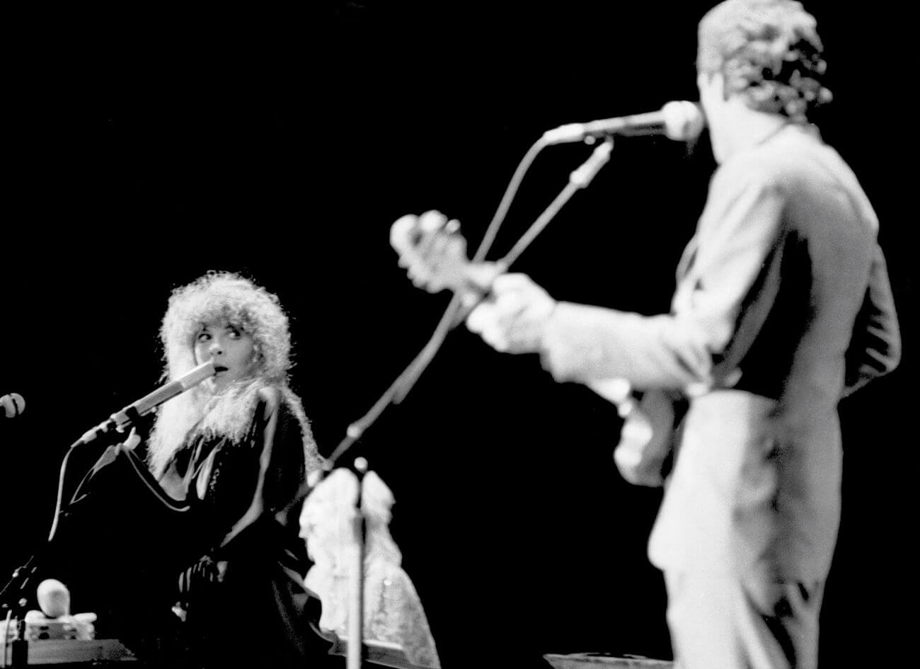 A black and white picture of Fleetwood Mac's Stevie Nicks singing into a microphone and looking at Lindsey Buckingham, who plays guitar and sings. 