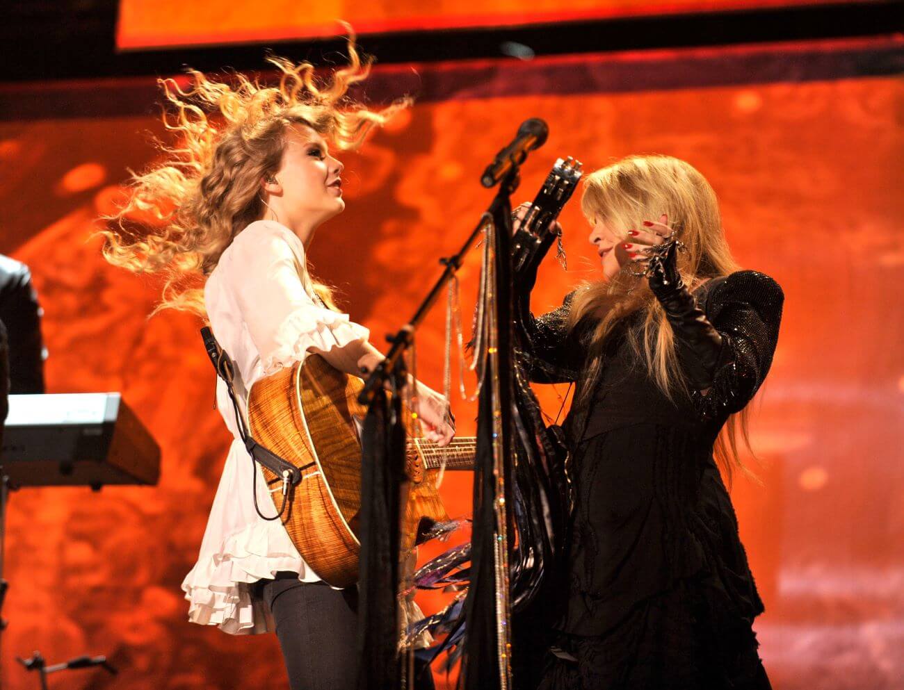 Taylor Swift plays guitar and stands on stage with Stevie Nicks, who has a tambourine. 