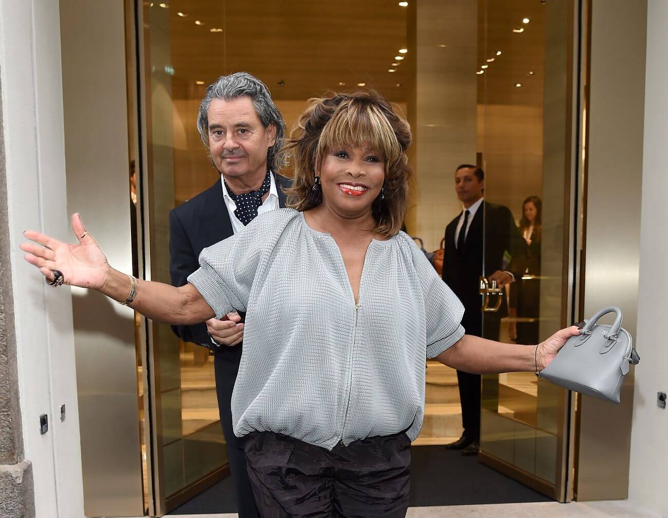 Tina Turner wears silver and holds a matching purse while her husband, Erwin Bach stands behind her.