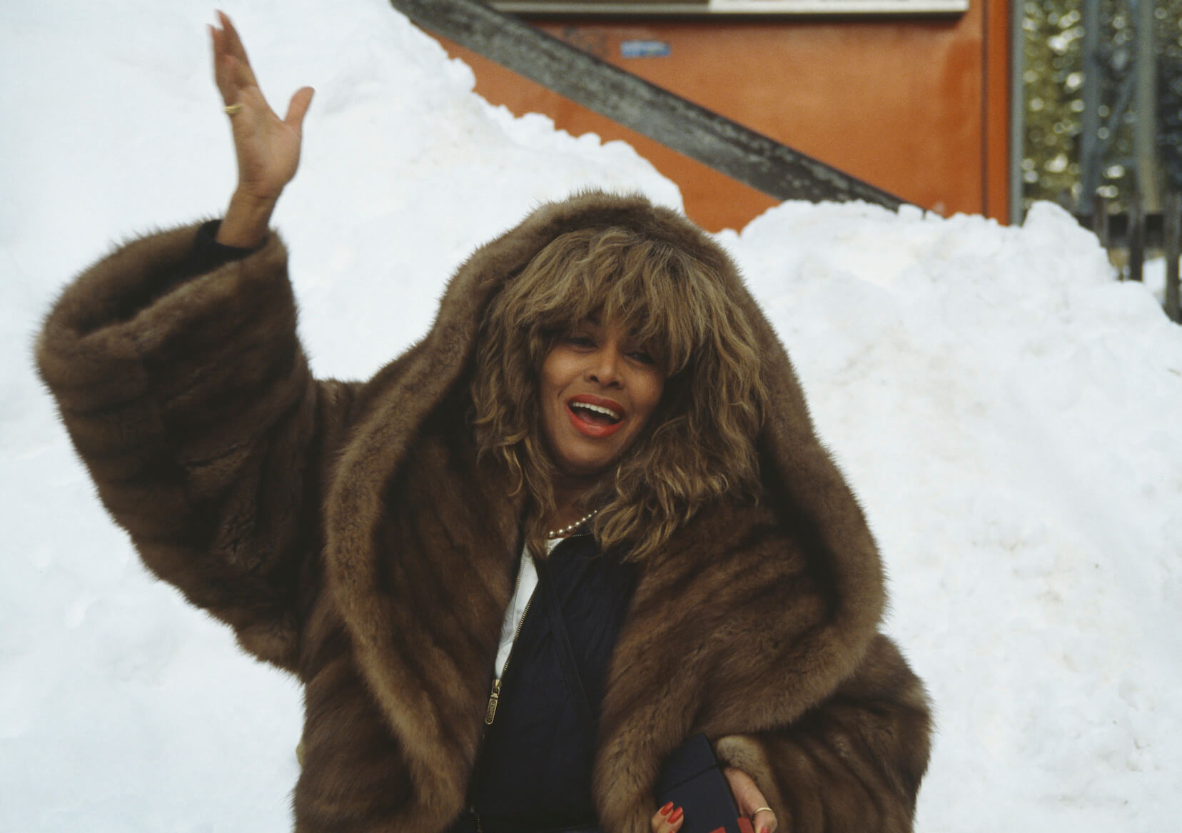 Tina Turner wearing a heavy coat outdoors in the snow while holding her hand up