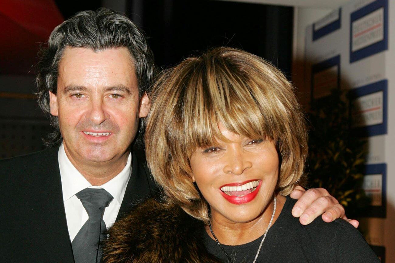 Erwin Bach, Tina Turner's husband, stands with his hand on her shoulder. They both smile.