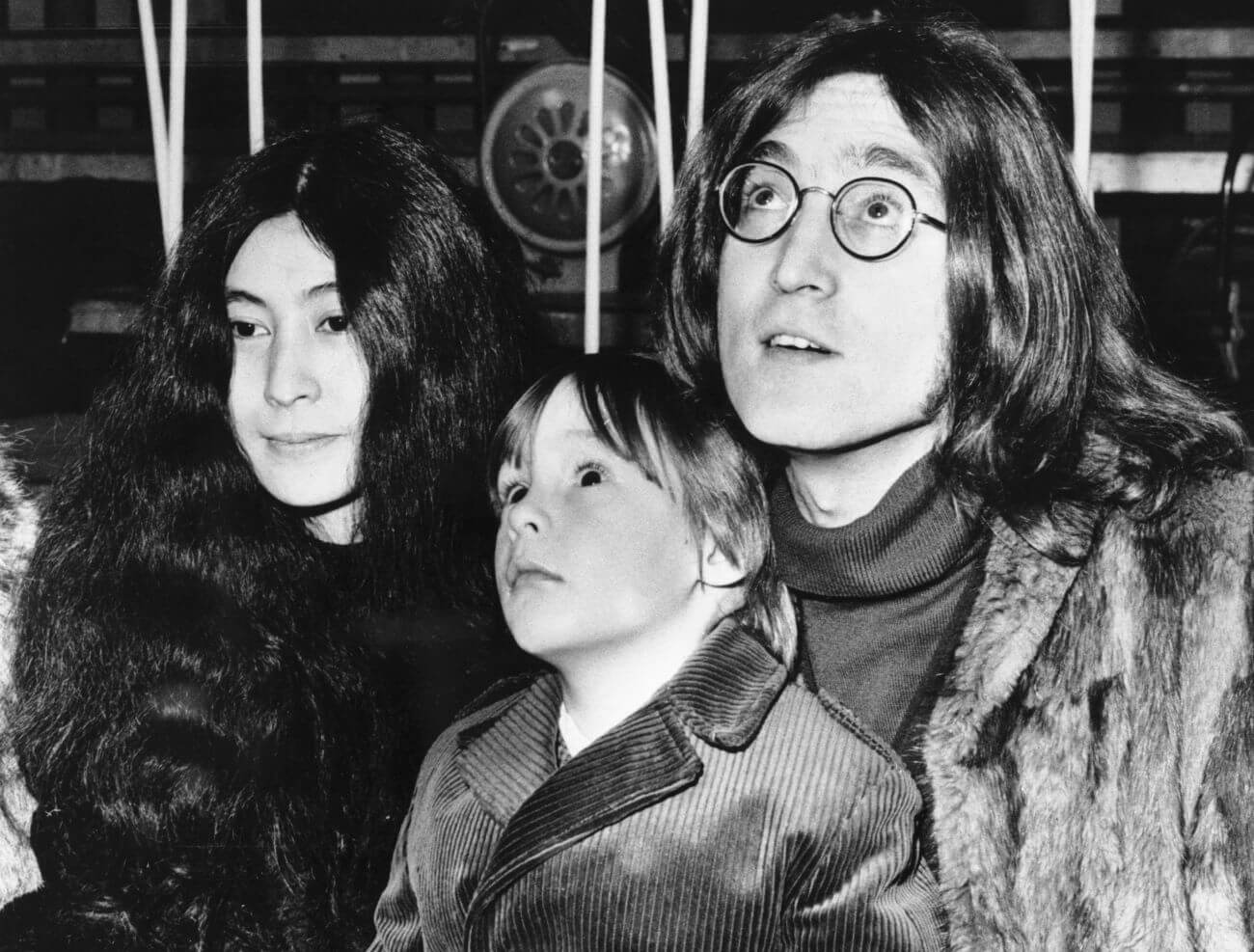 A black and white picture of Yoko Ono, Julian Lennon, and John Lennon. They all wear coats, and Julian sits on Lennon's lap.