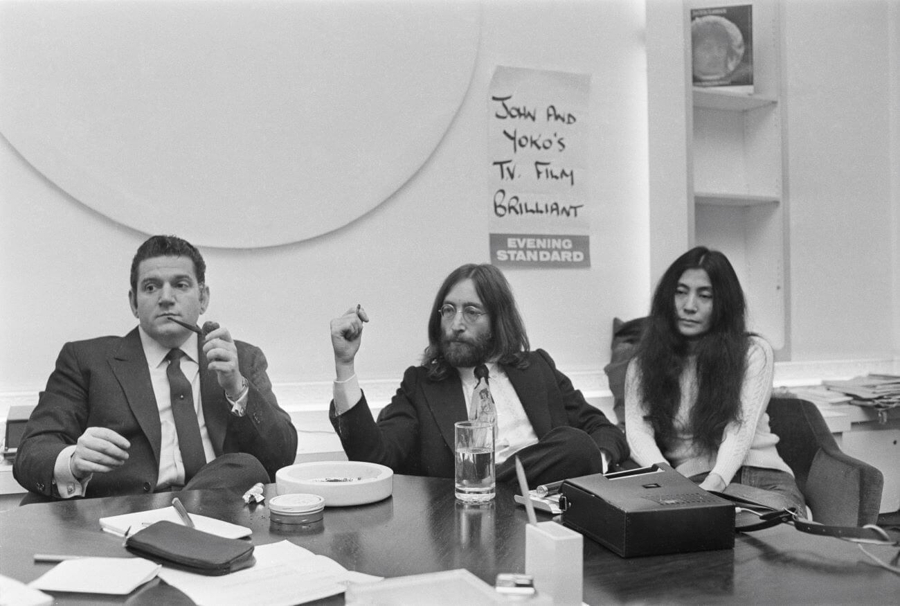 A black and white picture of Allen Klein, John Lennon, and Yoko Ono sitting at a table in front of a poster that says "John and Yoko's TV Film Brilliant."