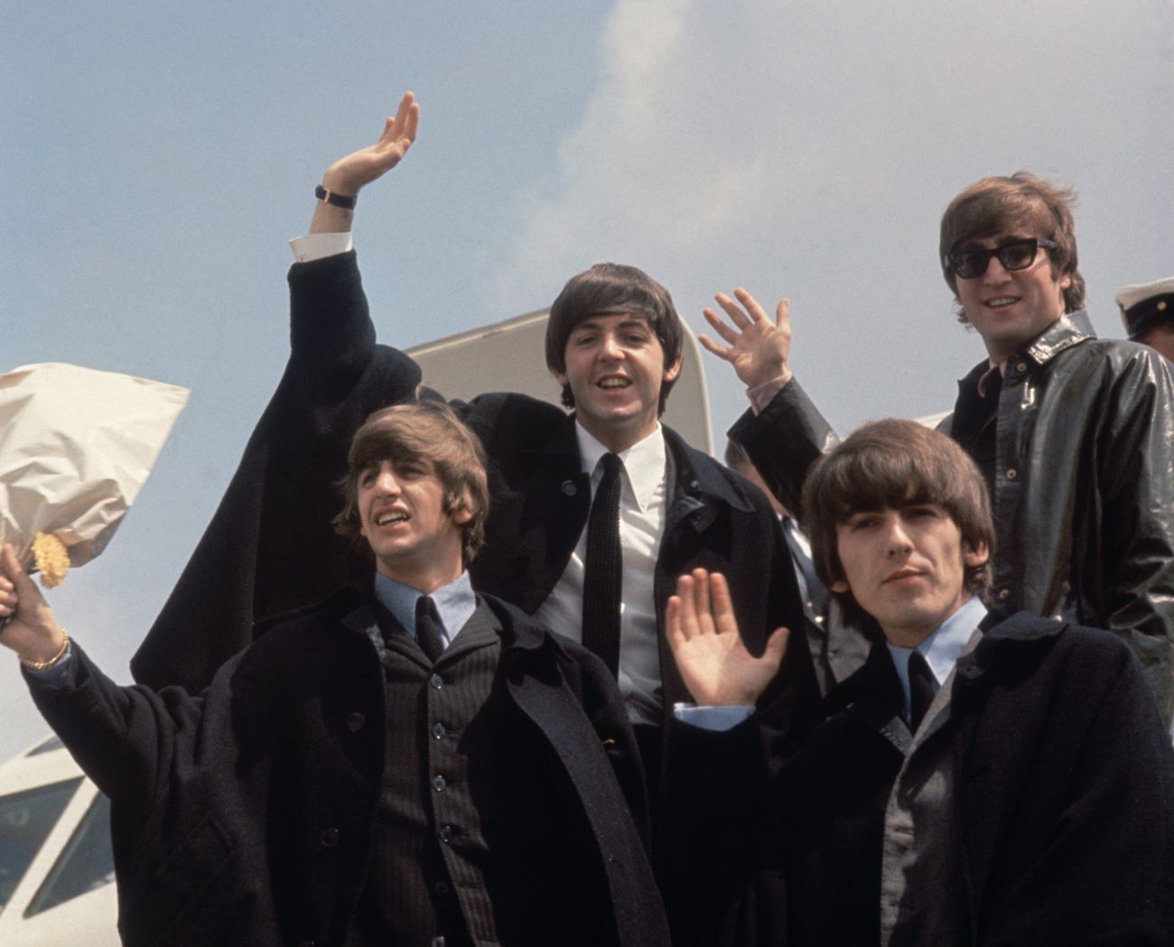 Ringo Starr, Paul McCartney, George Harrison, and John Lennon wave from the entrance to a plane. 