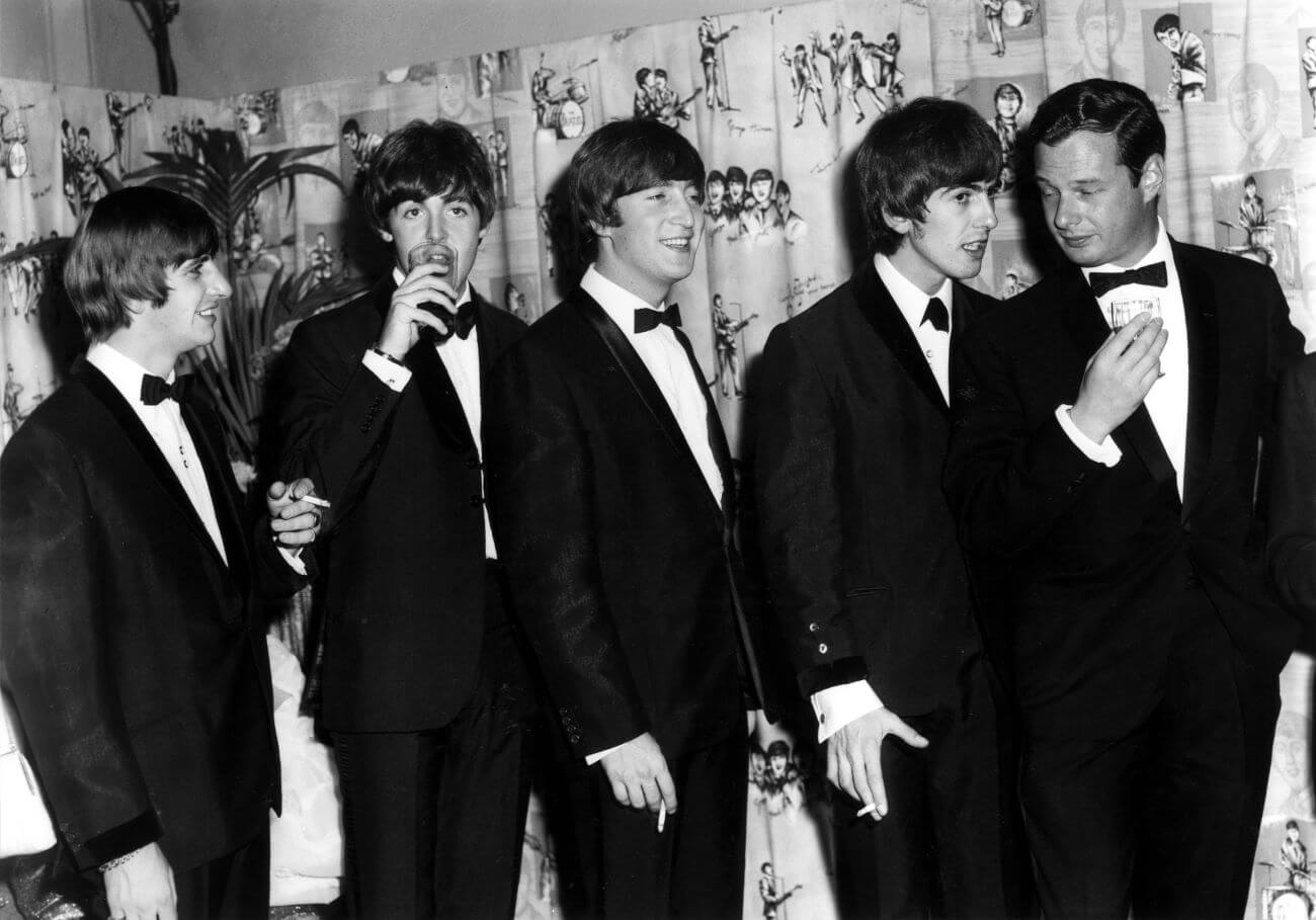 A black and white picture of Ringo Starr, Paul McCartney, John Lennon, and George Harrison standing in a line behind Brian Epstein. McCartney and Epstein hold drinks while Starr, Lennon, and Harrison hold cigarettes.