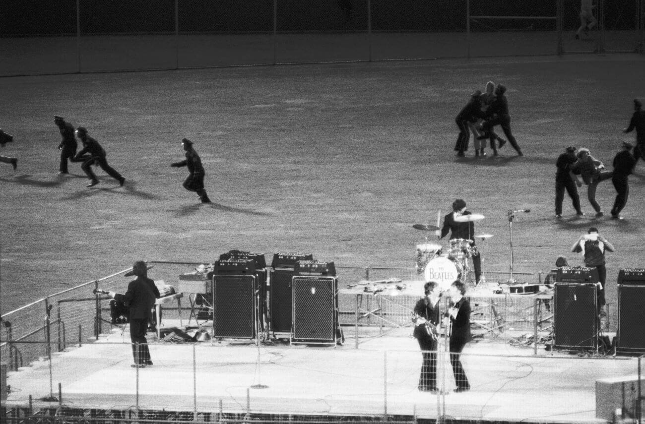 A black and white picture of The Beatles playing onstage while police officers arrest people.