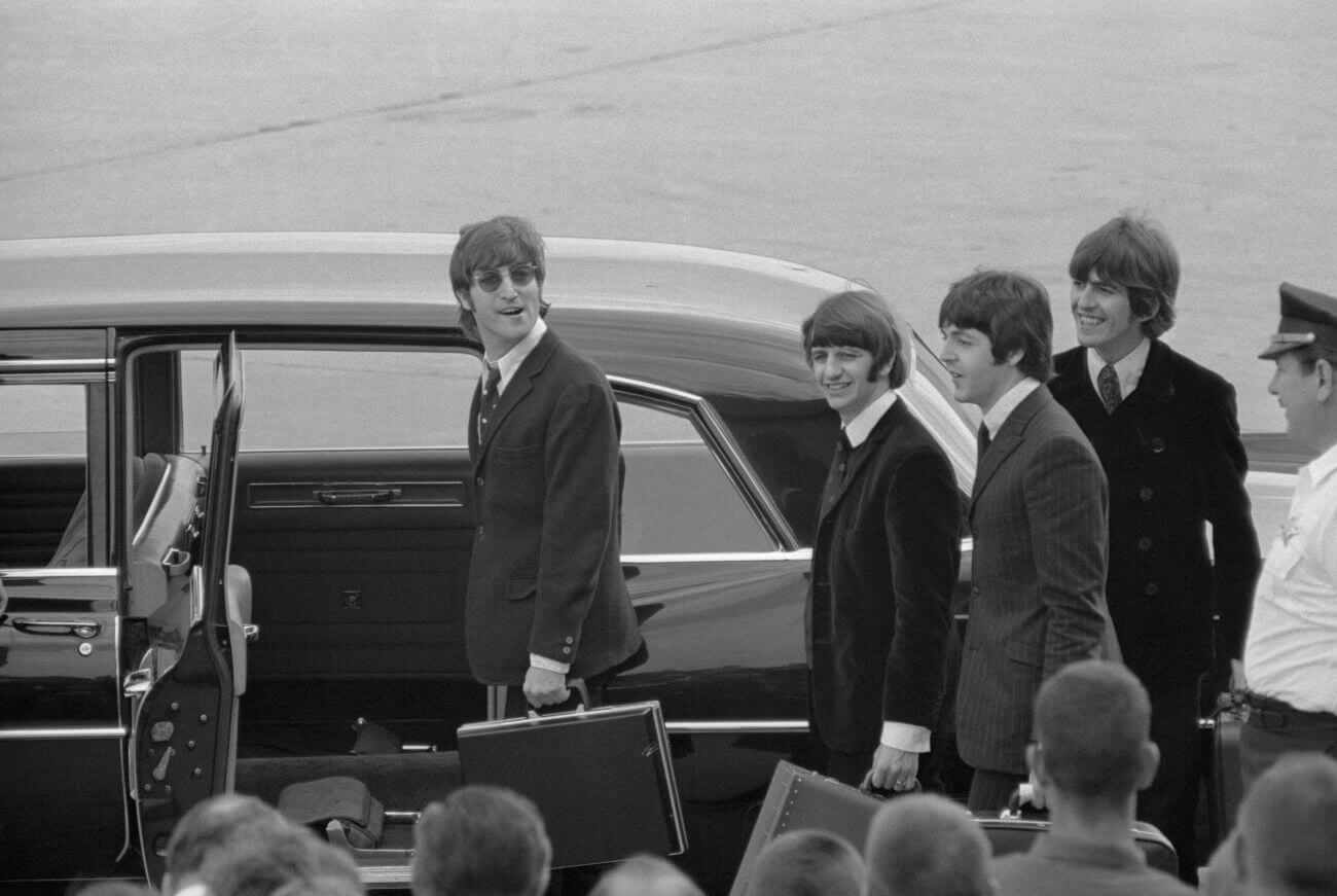 A black and white picture of John Lennon, Ringo Starr, George Harrison, and Paul McCartney holding suitcases and walking into a car.
