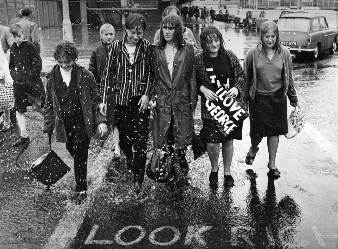 A black and white picture of Beatles fans walking through the rain. One holds a sign that says "I Love George."