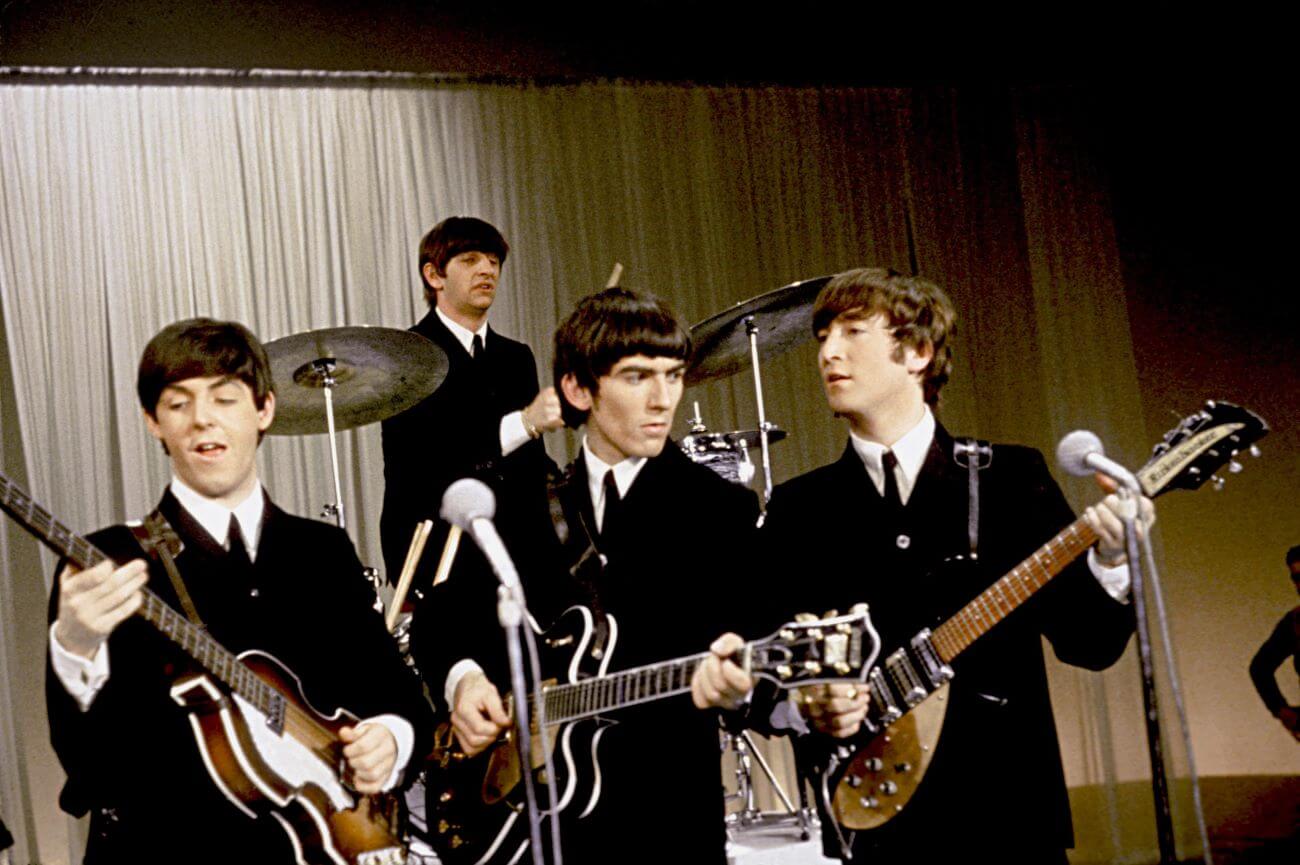 Paul McCartney, George Harrison, and John Lennon play guitars while Ringo Starr drums behind them.