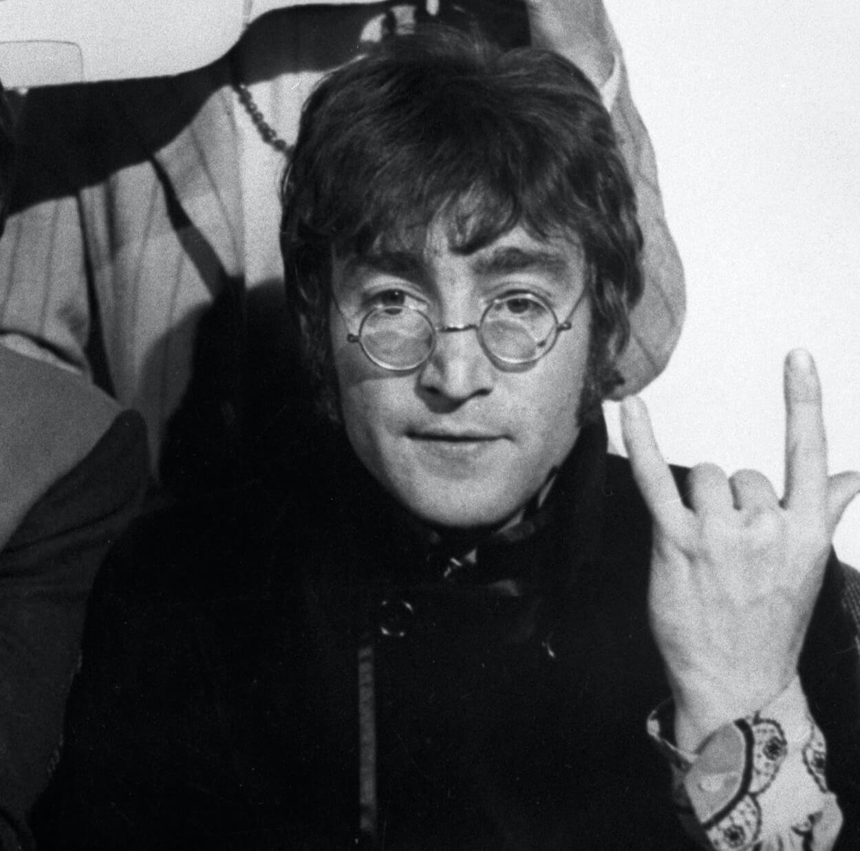 John Lennon wearing round glasses and extending the index finger and pinkie of his left hand into the air circa 1967.