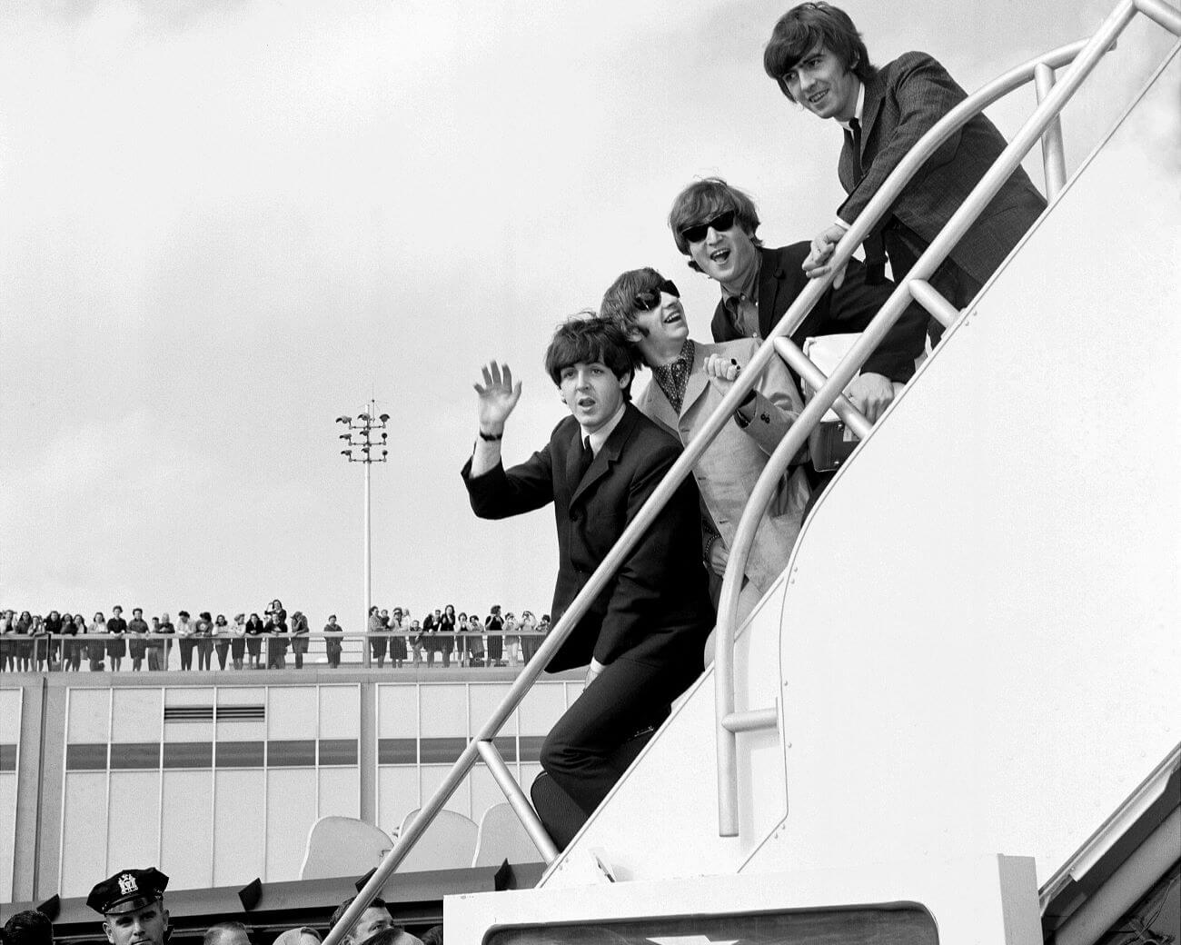 The Beatles stand on a stairway at an airport and wave to fans. 