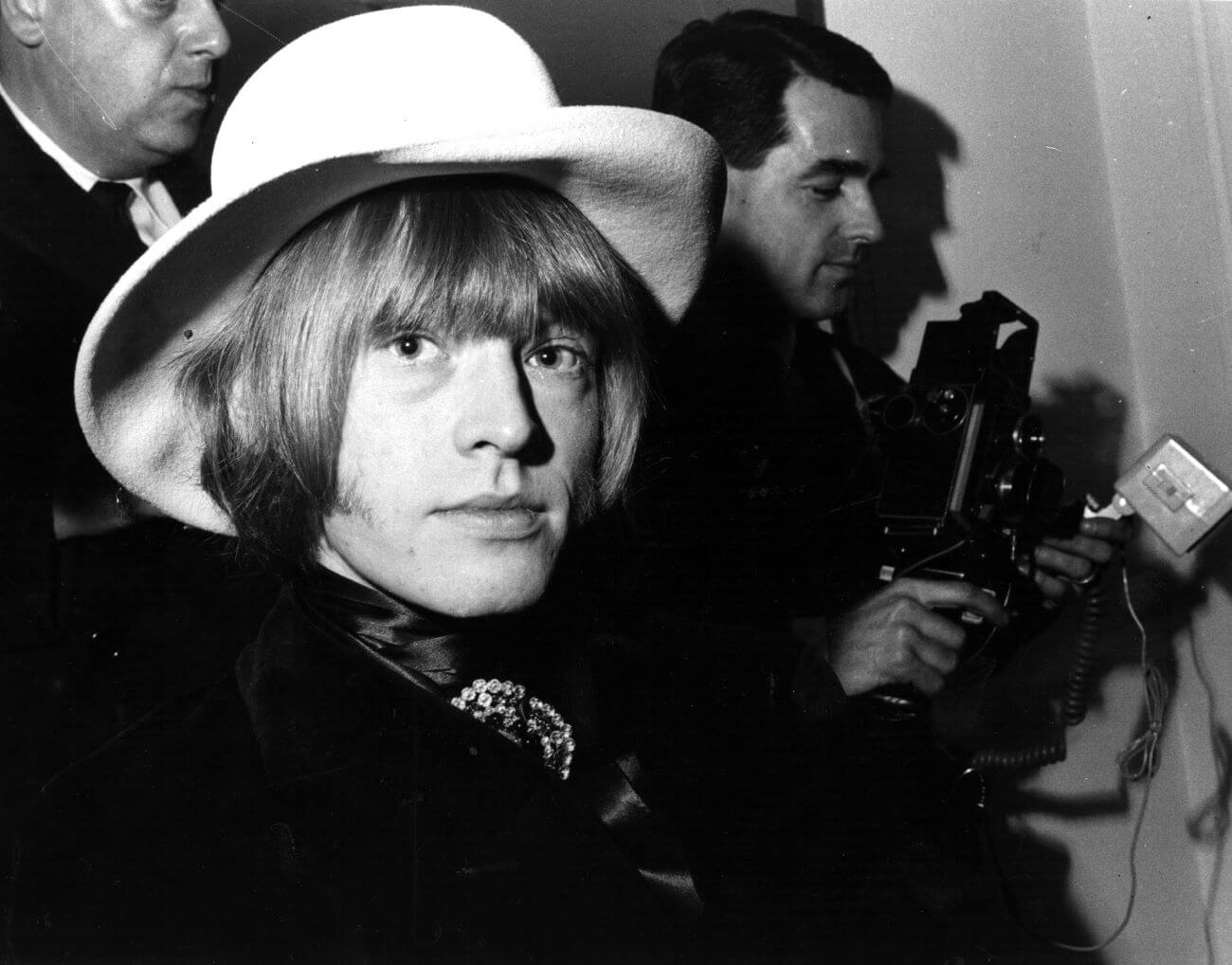 A black and white picture of Brian Jones wearing a hat in front of photographers.
