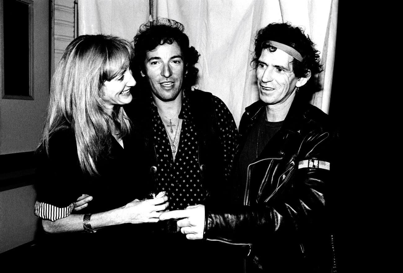A black and white picture of Patti Scialfa, Bruce Springsteen, and Keith Richards standing in front of a curtain with their arms around each other.