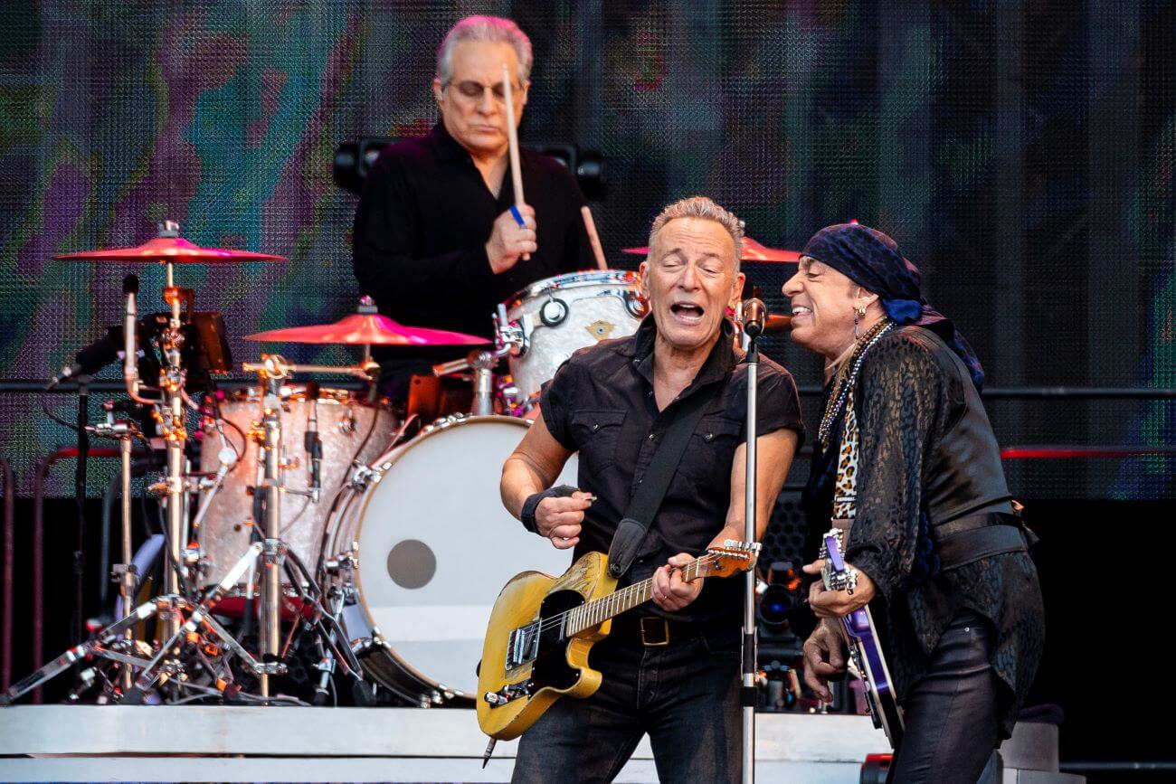 Bruce Springsteen and Steven Van Zandt sing into the same microphone while playing guitars. Max Weinberg drums on a platform behind them.