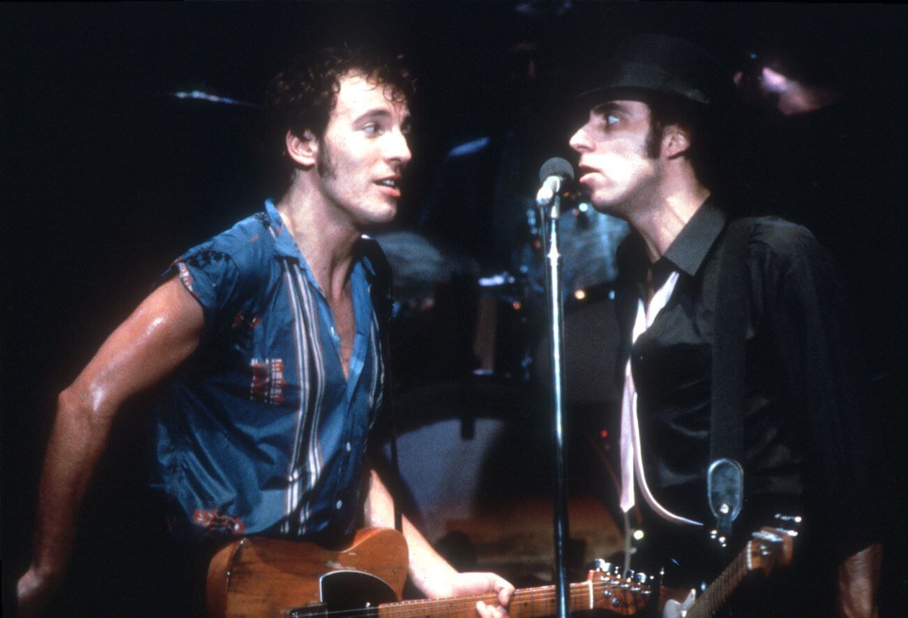 Bruce Springsteen holds a guitar and sings into the same microphone as Steven Van Zandt.