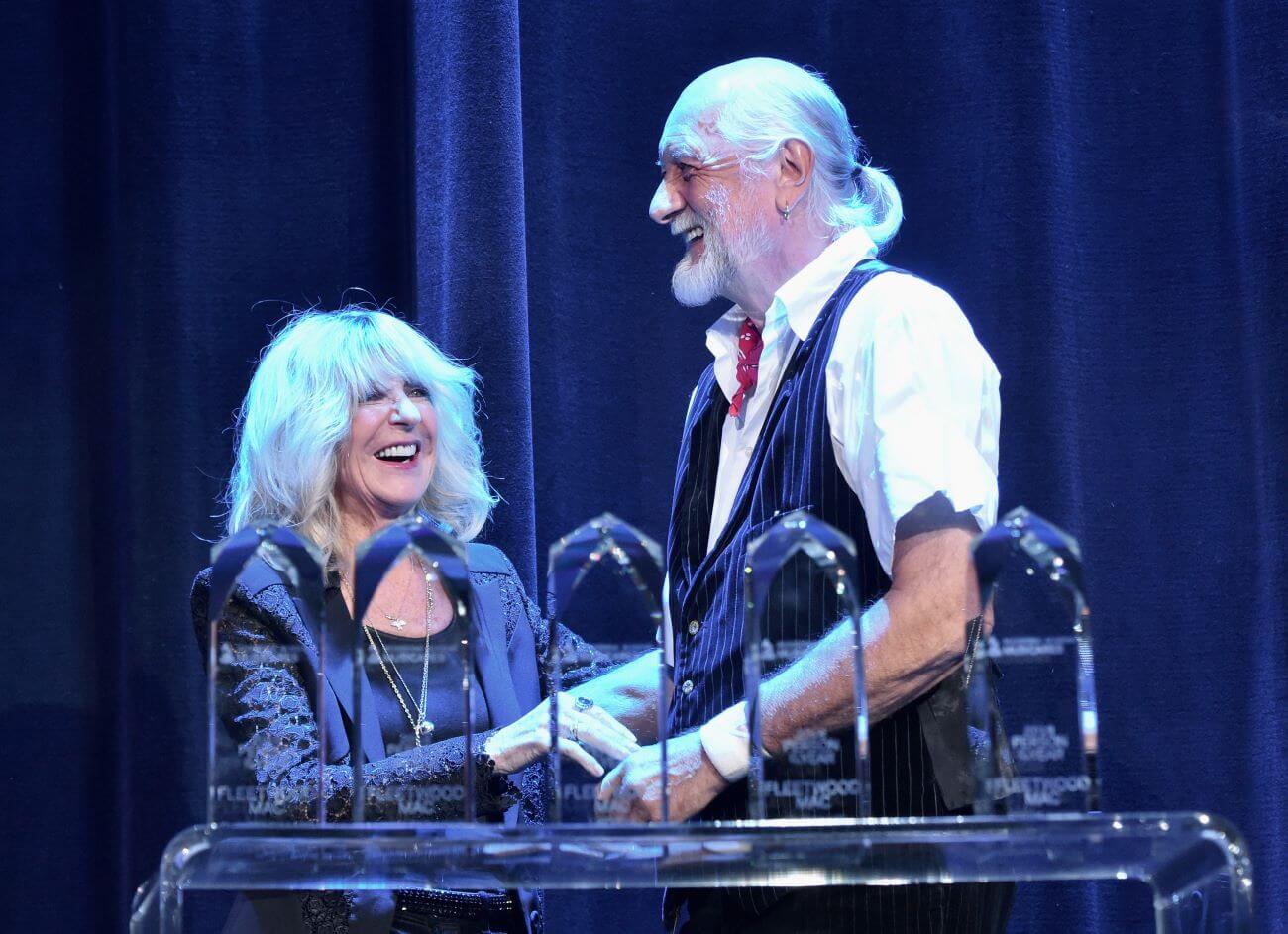 Christine McVie and Mick Fleetwood stand behind a podium laughing together.