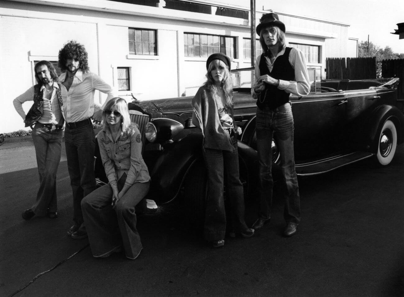 A black and white picture of John McVie, Lindsey Buckingham, Christine McVie, Stevie Nicks, and Mick Fleetwood posing around a car.