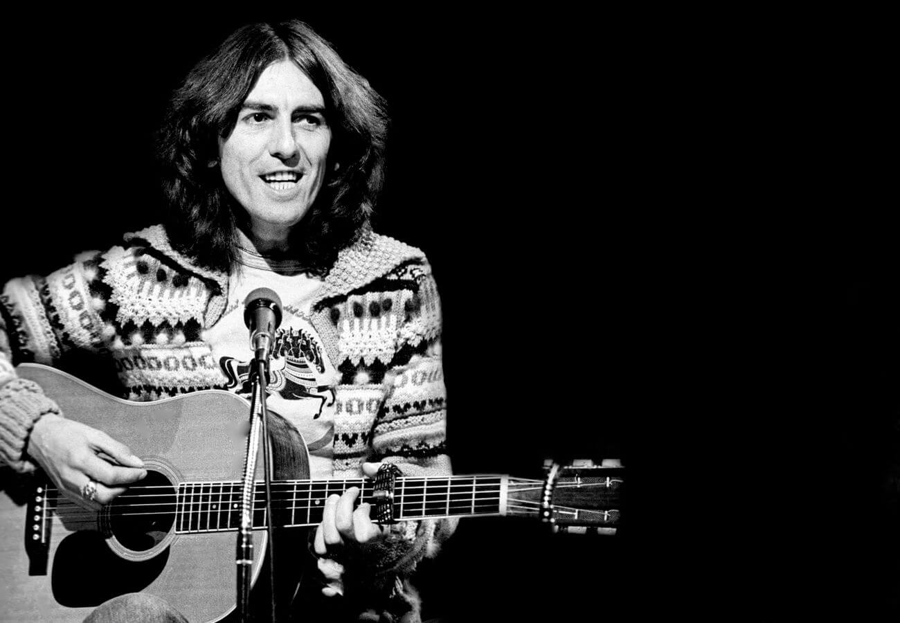 A black and white picture of George Harrison playing the acoustic guitar and wearing a sweater.