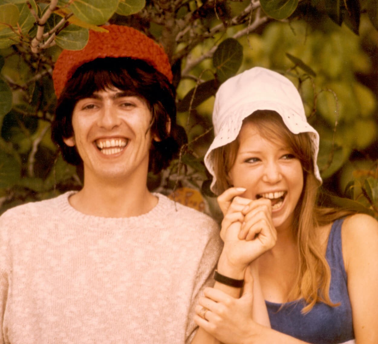 Beatles guitarist George Harrison (left) wearing a red hat and t-shirt and holding hands with his wife Pattie Boyd, wearing a white hat and blue top.